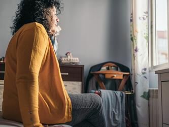 Older woman in yellow cardigan, sitting on bed, looking out of bedroom window